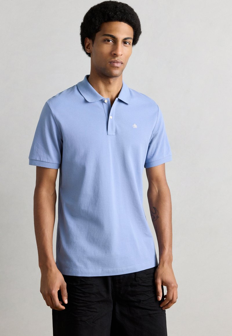 Ανοιχτό μπλε πόλο πουκάμισο από βαμβάκι, με μυτερό γιακά, κοντά μανίκια και λογότυπο στο αριστερό στήθος. Συνδυασμένο με μαύρο σορτς.