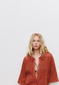 Rostfärgad stickad skjorta med krage, korta ärmar och öppen framsida, med texturerade mönster och dekorativa detaljer.