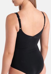 Mulher vista de costas a usar um fato de banho preto de uma peça com alças ajustáveis sobre um fundo liso.