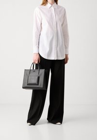 Persona con camicia bianca e pantaloni neri a gamba larga che tiene una borsa a spalla nera con tasca frontale texturizzata.