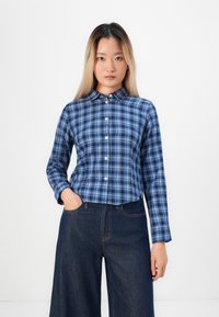 Kvinne med langt hår iført en blå rutete skjorte med knapper og mørke vide jeans, stående mot en hvit bakgrunn.