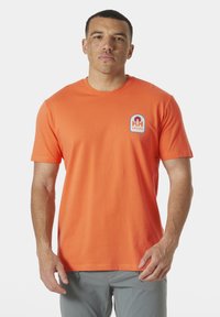 Krátký rukáv, tričko s kulatým výstřihem ve světlé oranžové barvě z bavlny, se modro-bílou nášivkou loga na levé hrudi. Hladká textura.