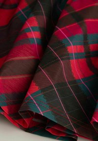 Czerwono-zielona tkanina w kratę z ciemnobrązowymi akcentami. Charakteryzuje się gładką fakturą oraz falistym wzorem wzdłuż krawędzi.