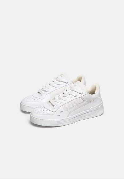 Coppia di sneakers bianche basse con punte perforate, pannelli laterali texturizzati e lacci bianchi su uno sfondo chiaro e semplice.