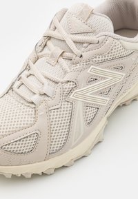 Beige színű sportcipő, hálós felső résszel, textúrázott részletekkel, fűzőkkel és mintázott talppal. Az oldalán jól látható "N" logó található.
