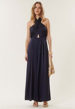 Femme aux cheveux blonds ondulés portant une combinaison sans manches de couleur bleu marine à col halter, tenant un sac beige à détails circulaires, debout pieds nus.