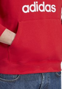 Osoba v červené mikině Adidas s bílým logem, ruka v přední kapse, v kombinaci s modrými džínami.