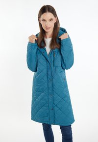 Teal quiltet lang frakk med hætte, der har en lynlåslomme og trykknaplukninger, parret med en hvid top og blå jeans.