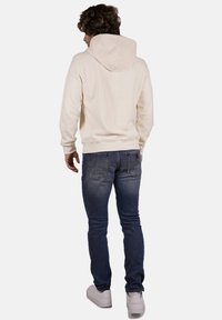 Felpa chiara con cappuccio e cordino, abbinata a jeans blu scuri e sneaker bianche, vista da dietro.