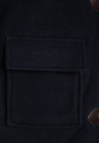 Cappotto di lana blu scuro con una grande tasca quadrata e un accento in pelle marrone vicino al bottone. Tessuto morbido con un design strutturato.