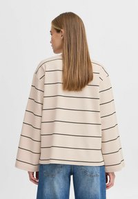 Sweatshirt beige à rayures avec manches longues, présentant des rayures noires horizontales et une coupe décontractée. Associé à un jean en denim bleu.