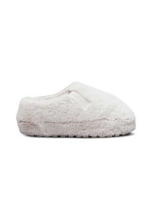 Zapatilla suave y de color beige claro, peluda, con puntera cerrada y suela plana, con una pequeña etiqueta de tela en el lateral.