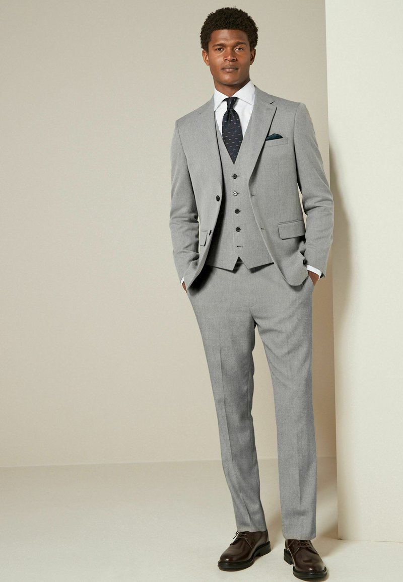 Light grey three-piece suit with a notched lapel, single-breasted jacket, matching waistcoat, and flat-front trousers. Dark shoes accent the outfit.