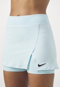 Falda deportiva de color azul claro con diseño de apertura, cinturilla elástica y pantalones cortos interiores. Presenta un logo negro de Nike en el lado inferior izquierdo.
