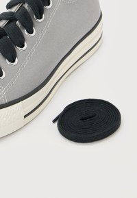Zapatilla de lona gris con cordones negros, suela de goma blanca, junto a un cordón negro enrollado sobre un fondo blanco.