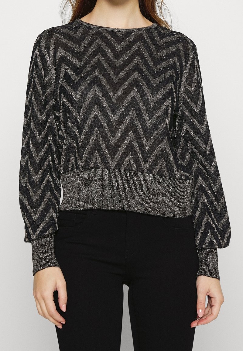 Femme portant un pull à manches longues noir et argenté à motif chevrons, associé à un pantalon noir uni, debout devant un fond blanc.