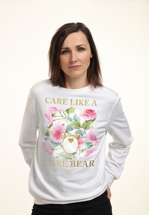 Hvid sweatshirt med en floral illustration, et stiliseret bjørneansigt og teksten "CARE LIKE A CARE BEAR" i guld, med 1982 nedenunder.