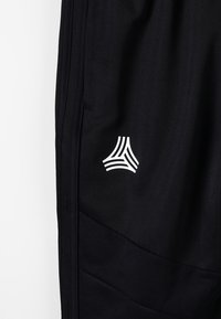 Schwarze Sporthosen aus strukturiertem Stoff, mit seitlichen Taschen und einem weißen Logo mit geschwungenen Linien am Unterschenkel.