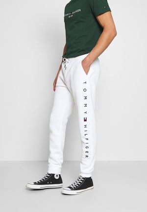 Person in weißen Tommy Hilfiger Jogginghosen mit schwarzen High-Top Converse Sneakern und einem dunklen grünen Tommy Hilfiger T-Shirt, der an einer weißen Wand steht.
