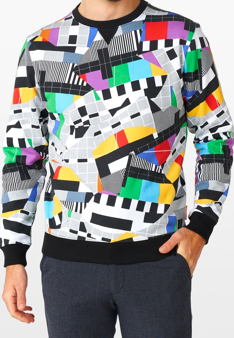 Homme portant un sweat-shirt à manches longues avec un motif géométrique et rayé coloré, associé à un pantalon texturé foncé.