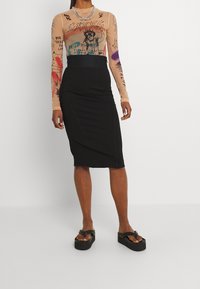 Femme portant un haut graphique transparent à manches longues, une jupe crayon noire taille haute et des sandales plateformes noires contre un fond uni.