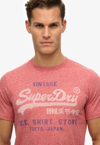 Camiseta roja de manga corta, fabricada con un tejido texturizado, con texto impreso en azul y blanco desvaído que dice: "VINTAGE SuperDry" y otros detalles.