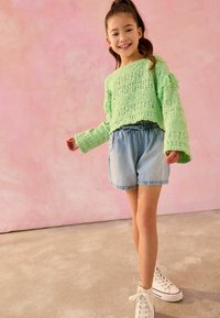 Hellgrüner Strickpullover mit Fransenärmeln, kombiniert mit Denim-Shorts und weißen High-Top-Sneakern auf einem hellgrauen Boden.