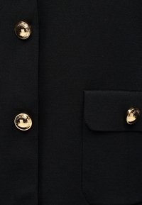 Μαύρο ύφασμα με τρία λαμπερά χρυσά κουμπιά, δύο σε ραφή και ένα σε ορθογώνιο πατιλέτα τσέπης. Τα κουμπιά έχουν χαραγμένο κείμενο.