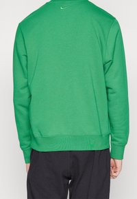 Grön Nike-tröja med långa ärmar, ribbade muddar och fåll. Har en liten swoosh-logga på övre ryggen. Mjuk tygtextur.