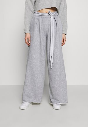 Person trägt graue Weitbein-Sweathose mit gebundenem Gürtel, gekürztes graues Sweatshirt und weiße Turnschuhe, steht vor einem einfarbigen Hintergrund.