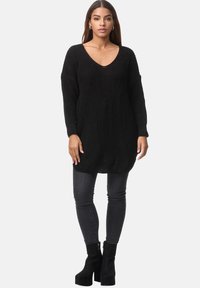 Schwarzer gestrickter Tunika-Pullover mit V-Ausschnitt, langen Ärmeln und lockerem Schnitt, kombiniert mit dunklen Skinny-Jeans und schwarzen, hochhackigen Ankle Boots.