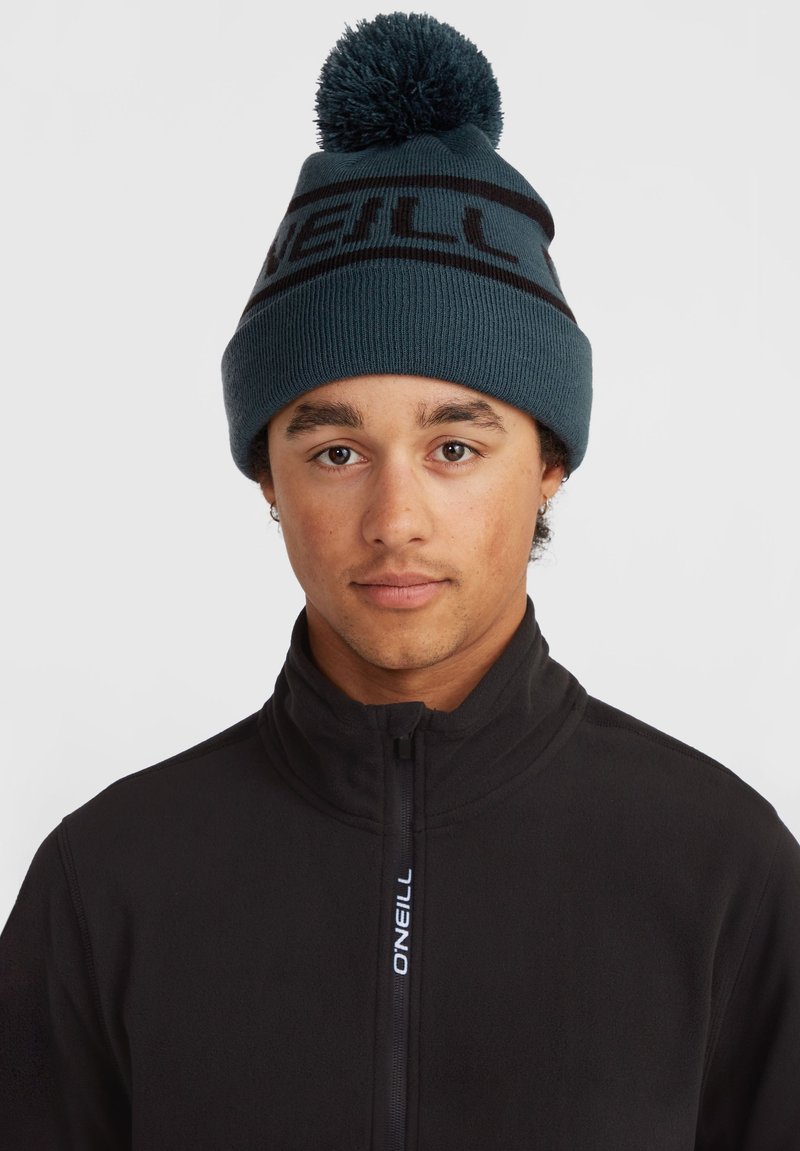 Teal knitted beanie with a black logo band and a pom-pom on top. Black fleece jacket with a high collar and zipper detail.
