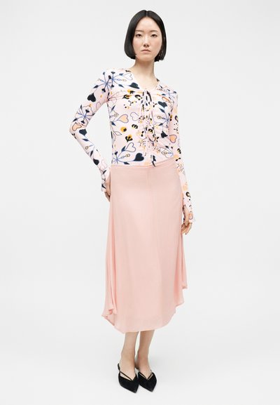 Femme aux cheveux courts noirs portant un haut à manches longues à motifs floraux avec un laçage sur le devant, une jupe midi fluide rose pâle et des chaussures noires à bout pointu.