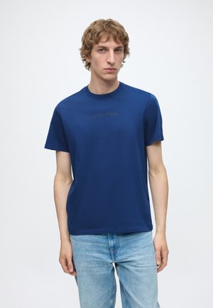 Ung mand med kort, krøllet hår iført en blå Calvin Klein t-shirt og lyseblå jeans mod en ensfarvet hvid baggrund.