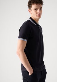 Crna polo majica s klasičnom kragnom, s naglascima svijetloplave i bijele pruge na manžetama. Napravljena od teksturirane tkanine.