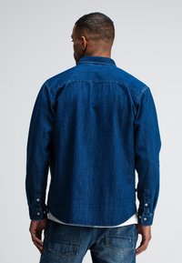 Donkerblauwe denim overhemd, met knoopmanchetten, rugdetail, lange mouwen en een gebogen zoom. De stof heeft een robuuste textuur met zichtbare naden.