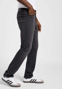 Jeans de mezclilla gris oscuro con un diseño de pierna recta. Presenta costuras claras, cinco bolsillos y un parche con logo en la cinturilla. Combinados con zapatillas blancas.