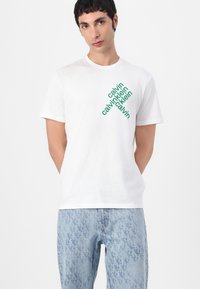 Bijela pamučna majica s zelenim dijagonalnim tekstom "calvin klein" na prednjoj strani, u kombinaciji s svijetloplavim trapericama s uzorkom.