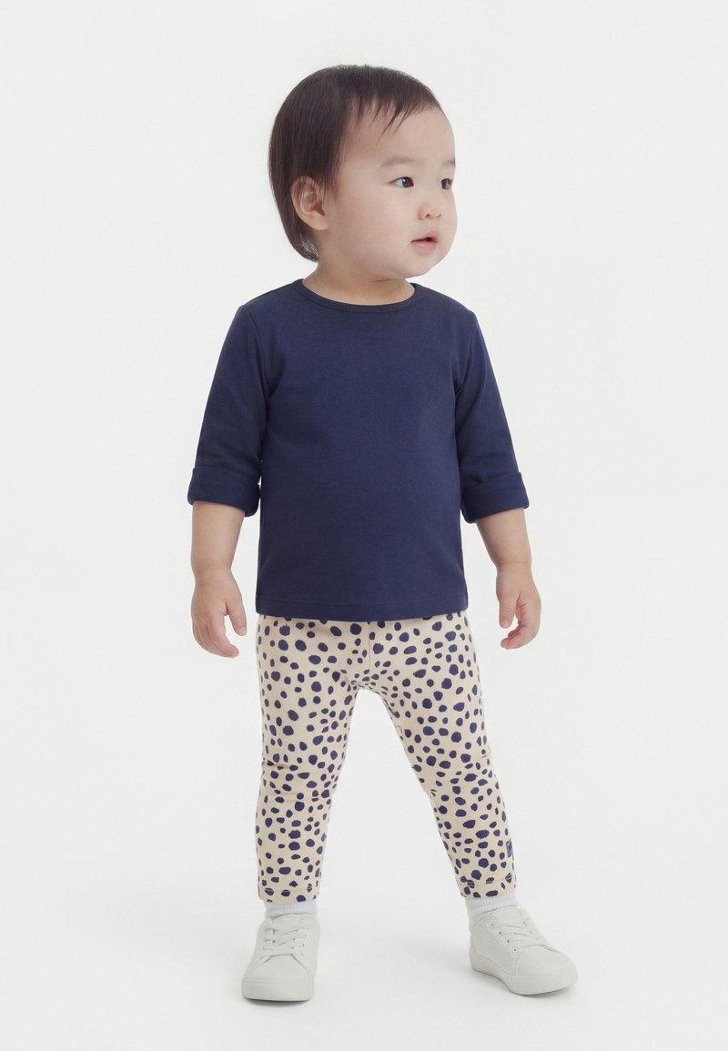 Μικρό παιδί όρθιο, φοράει μπλε μακρυμάνικη μπλούζα, μπεζ κολάν με μπλε πουά και λευκά αθλητικά, κοιτάζει προς τα δεξιά.