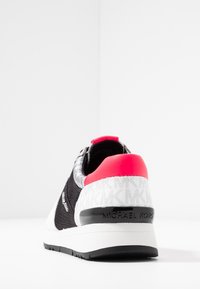 Svart mesh-sneaker med en vit och texturerad häl, med en rosa accent bak och "MICHAEL KORS"-logotyp i grått.