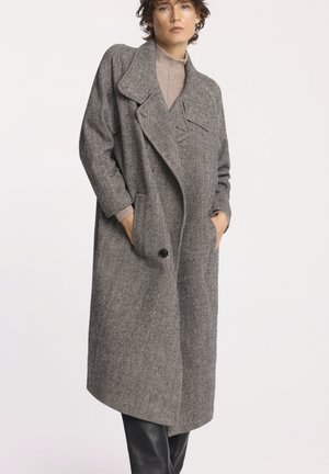 Kobieta ubrana w długi szary płaszcz w jodełkę, nosząca beżowy sweter z golfem i czarne spodnie, stoi z rękami w kieszeniach płaszcza.