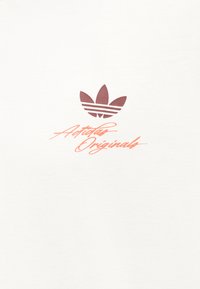 Adidas Originals logotipas su tamsiai raudonu trefoilio simboliu viršuje, kuriame geltonos spalvos kursyvinis užrašas „Adidas Originals“ ant baltos audinio fono.