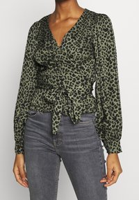 Blusa avvolgente in verde oliva con stampa leopardata nera, maniche lunghe a sbuffo, scollatura a V e dettaglio della vita annodata. Abbinata a jeans scuri.