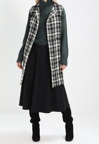 Czarna i biała kamizelka w kratę na zielonym, prążkowanym swetrze, zestawiona z czarną, luźno opadającą spódnicą i czarnymi kozakami za kolano. Gładkie tekstury i warstwowa konstrukcja.