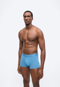 Μυώδης άνδρας όρθιος, φοράει ανοιχτό γαλάζιο μποξεράκι Calvin Klein, σε απλό φωτεινό γκρι φόντο, κοιτάζοντας μπροστά.