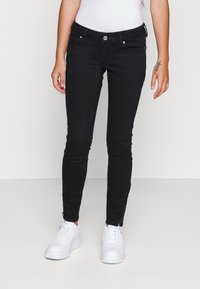 Persona vistiendo jeans negros ajustados con detalles de cremallera en los tobillos y zapatillas blancas, de pie contra un fondo liso claro.