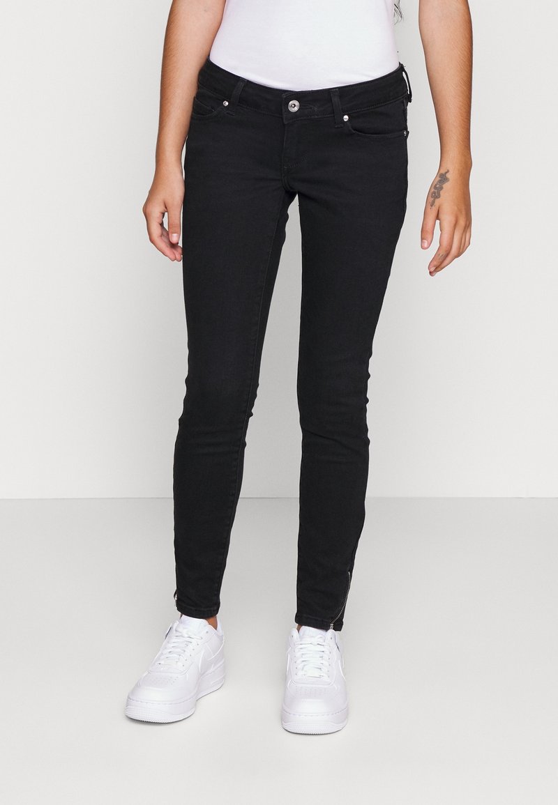 Persona vistiendo jeans negros ajustados con detalles de cremallera en los tobillos y zapatillas blancas, de pie contra un fondo liso claro.