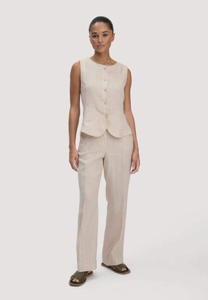 Vrouw staat en draagt een beige mouwloos topje met knopen en bijpassende wijde broekspijpen met donkere open-toe platte sandalen op een lichte achtergrond.