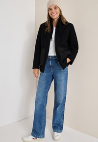 Giacca in pile nera con colletto alto e dettagli neri, abbinata a jeans blue chiaro a gamba larga e sneakers bianche. Berretto di maglia color crema.