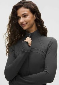 Mujer con cabello largo y rizado que lleva una chaqueta deportiva gris oscuro con cremallera, tirando de la cremallera cerca de su cuello, sonriendo suavemente.